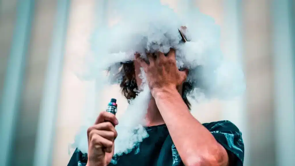 Person vaping with thick cloud of vapor covering face and holding a vape device