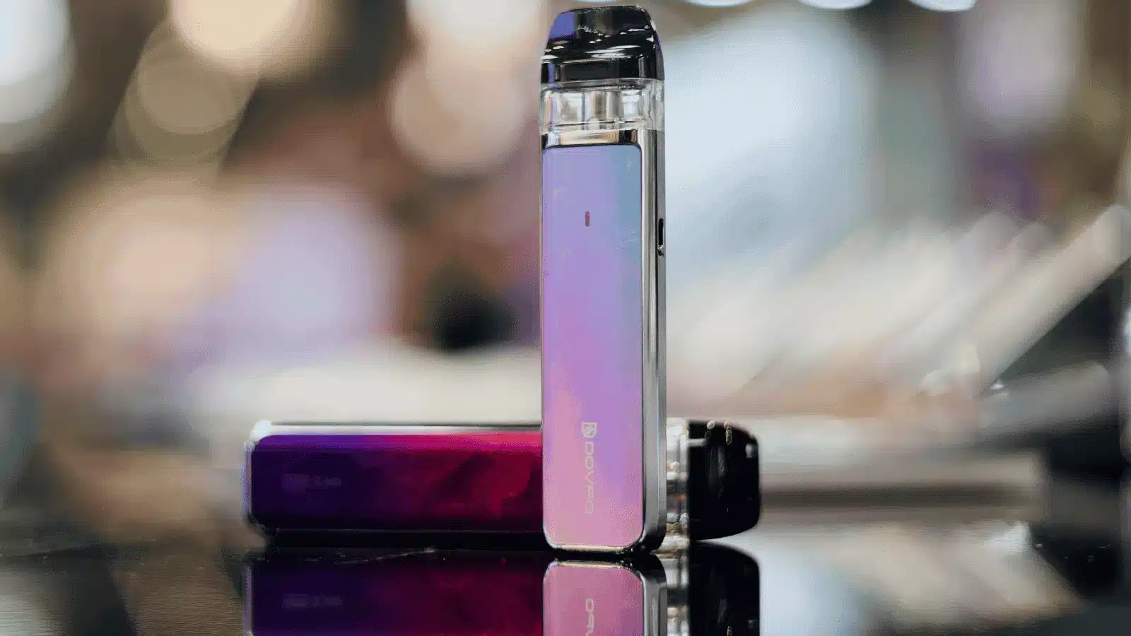 Close-up of a modern vape device with a sleek, purple-pink gradient design, displayed upright on a reflective surface with a blurred background of lights
